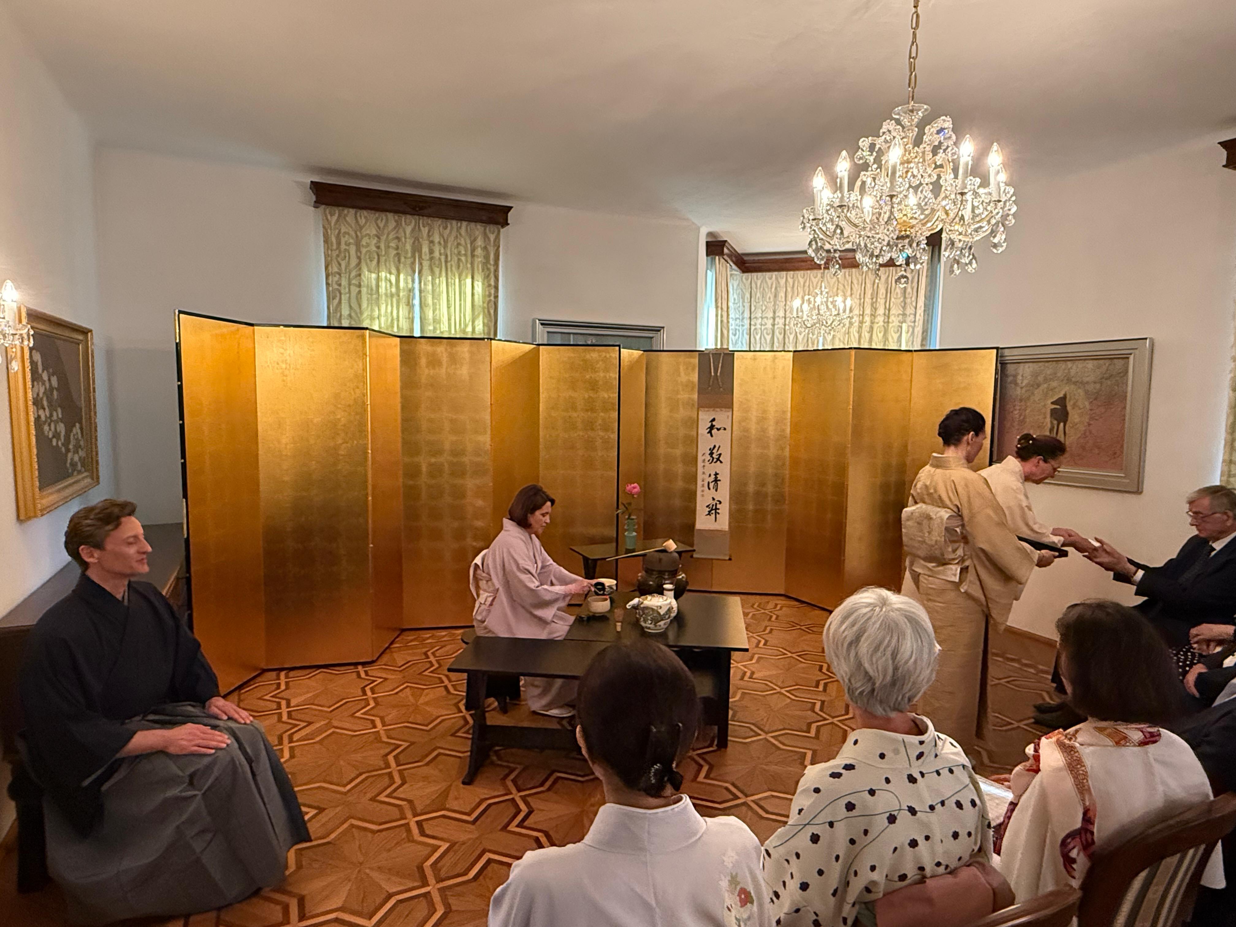 Formal Ryūrei tea gathering at the Residence of the Ambassador of Japan in Slovenia, with guests seated around the host preparing matcha before a gold folding screen and a scroll reading “Wa Kei Sei Jaku”.